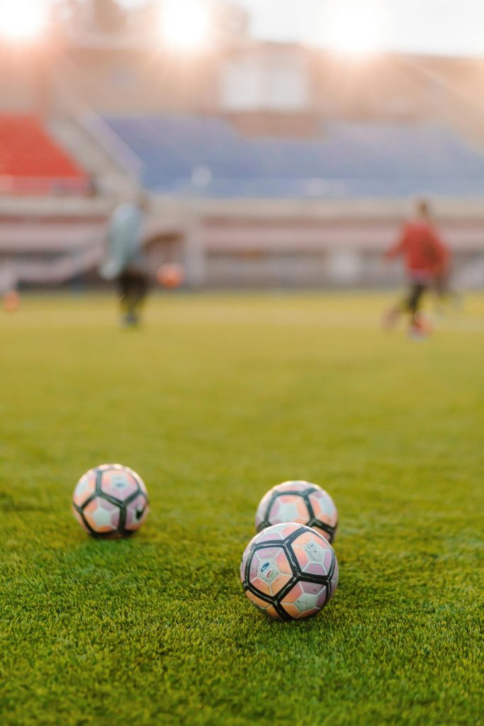 Trois ballons de football sur un terrain en herbe avec des joueurs flous s'entraînant en arrière-plan.